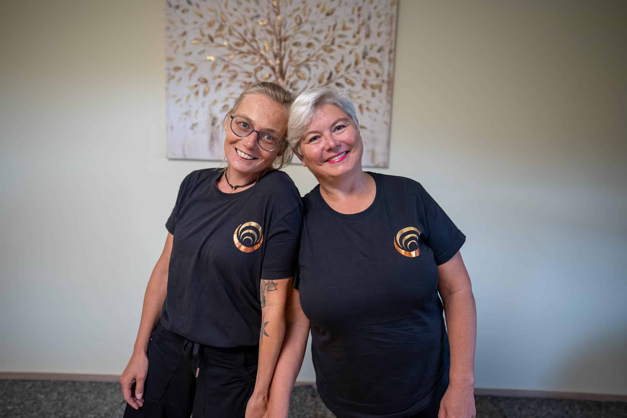 Zwei Personen in Shiatsu-Shirts lächeln in die Kamera, vor bildhaftem Hintergrund einer beruhigenden Atmosphäre.