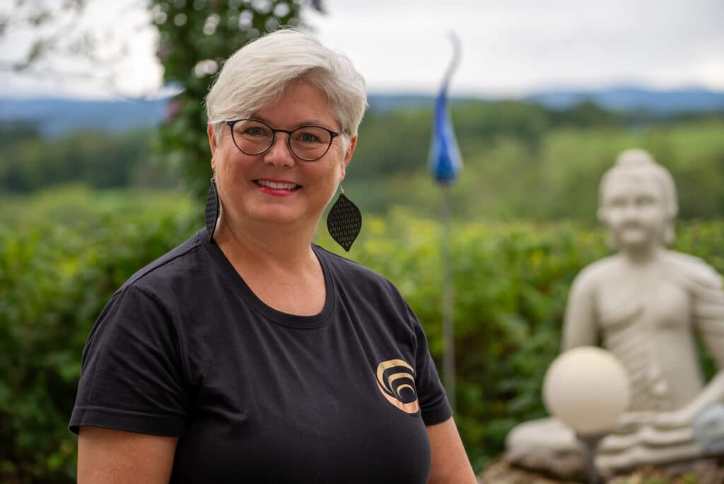 Eine Person lächelt im Freien, im Hintergrund ist eine Buddha-Statue und Landschaft zu sehen.