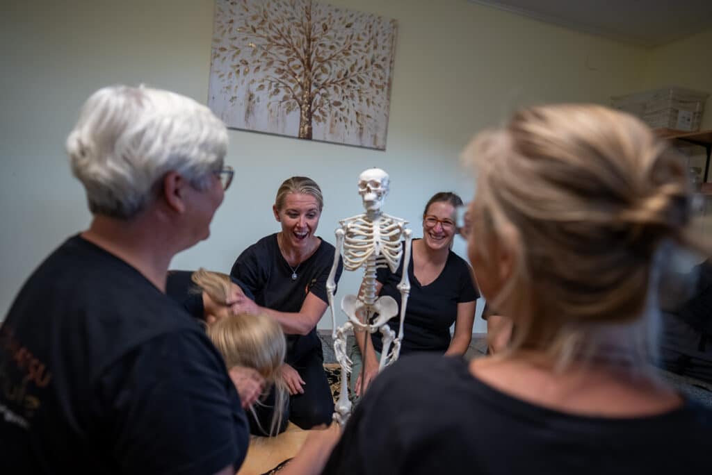 Personen in einem Shiatsu-Kurs lachen und interagieren mit einem Skelettmodell in einem hellen Raum.