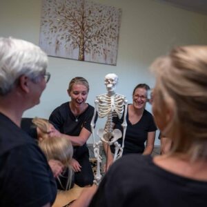 Personen in einem Shiatsu-Kurs lachen und interagieren mit einem Skelettmodell in einem hellen Raum.