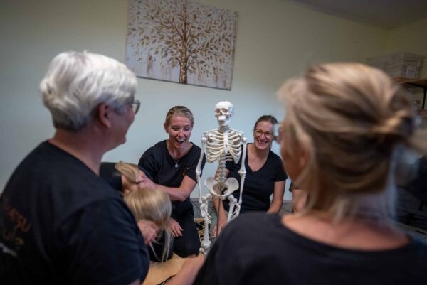 Personen in einem Shiatsu-Kurs lachen und interagieren mit einem Skelettmodell in einem hellen Raum.