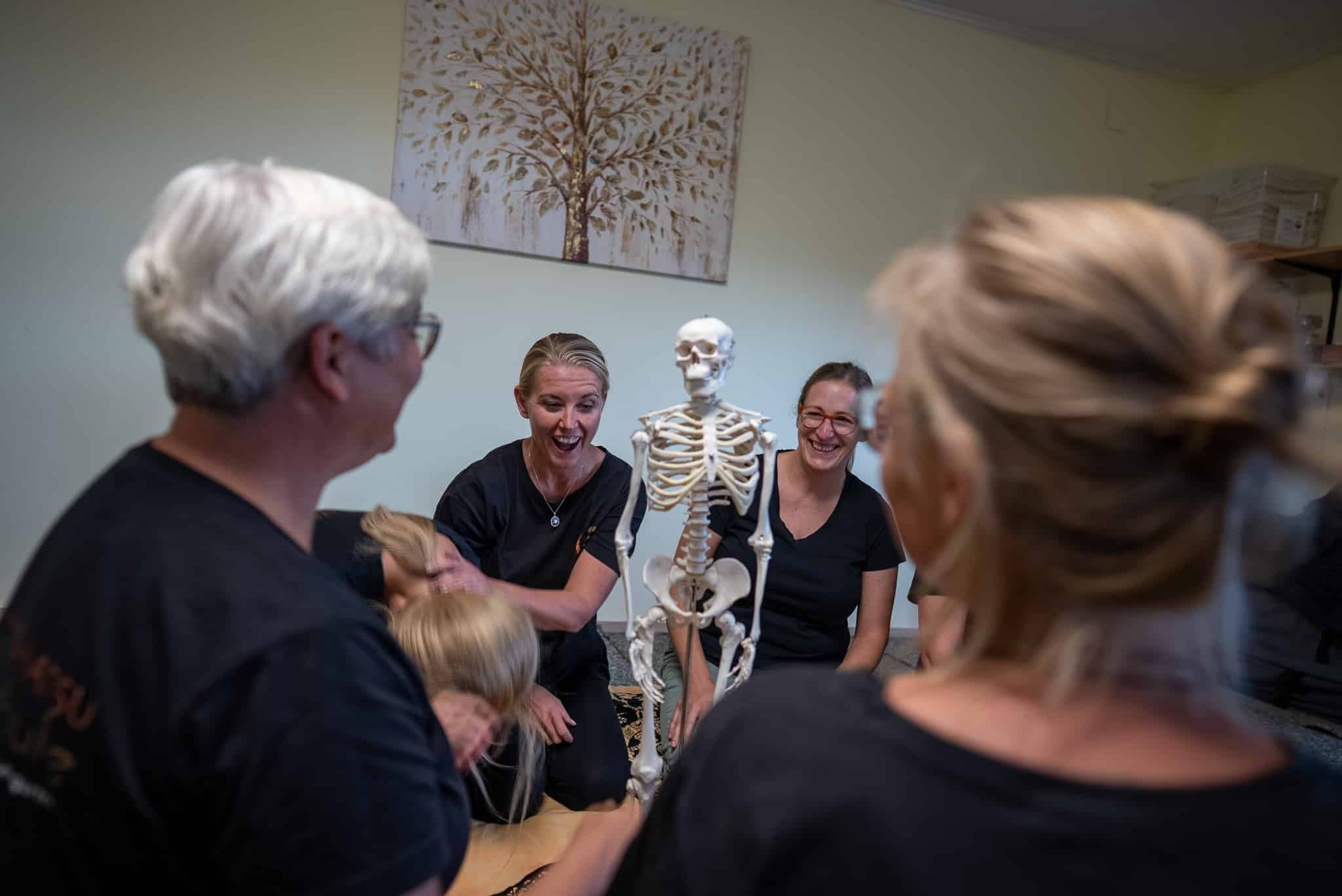 Personen in einem Shiatsu-Kurs lachen und interagieren mit einem Skelettmodell in einem hellen Raum.